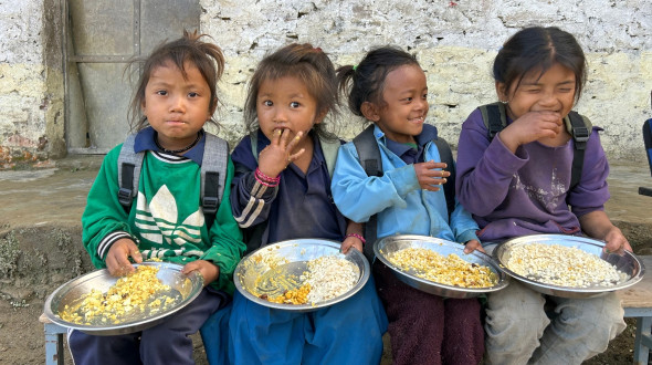 Vier Kinder sitzen auf einer Bank und essen ihre Schulmahlzeit (Reis mit Kichererbsen und hartgekochtem Ei)